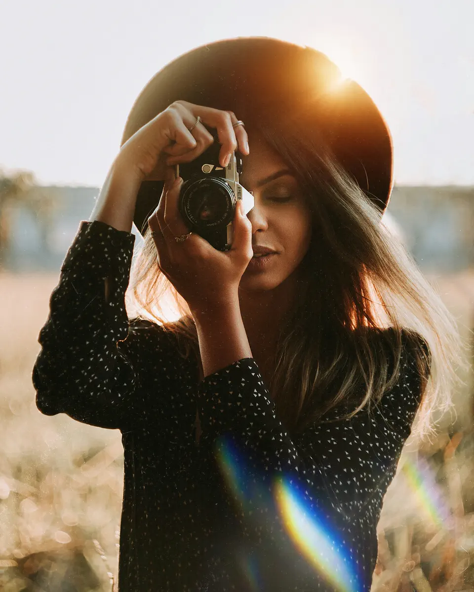 A photographer in a wide-brimmed hat raising a film camera to her eye in golden backlight