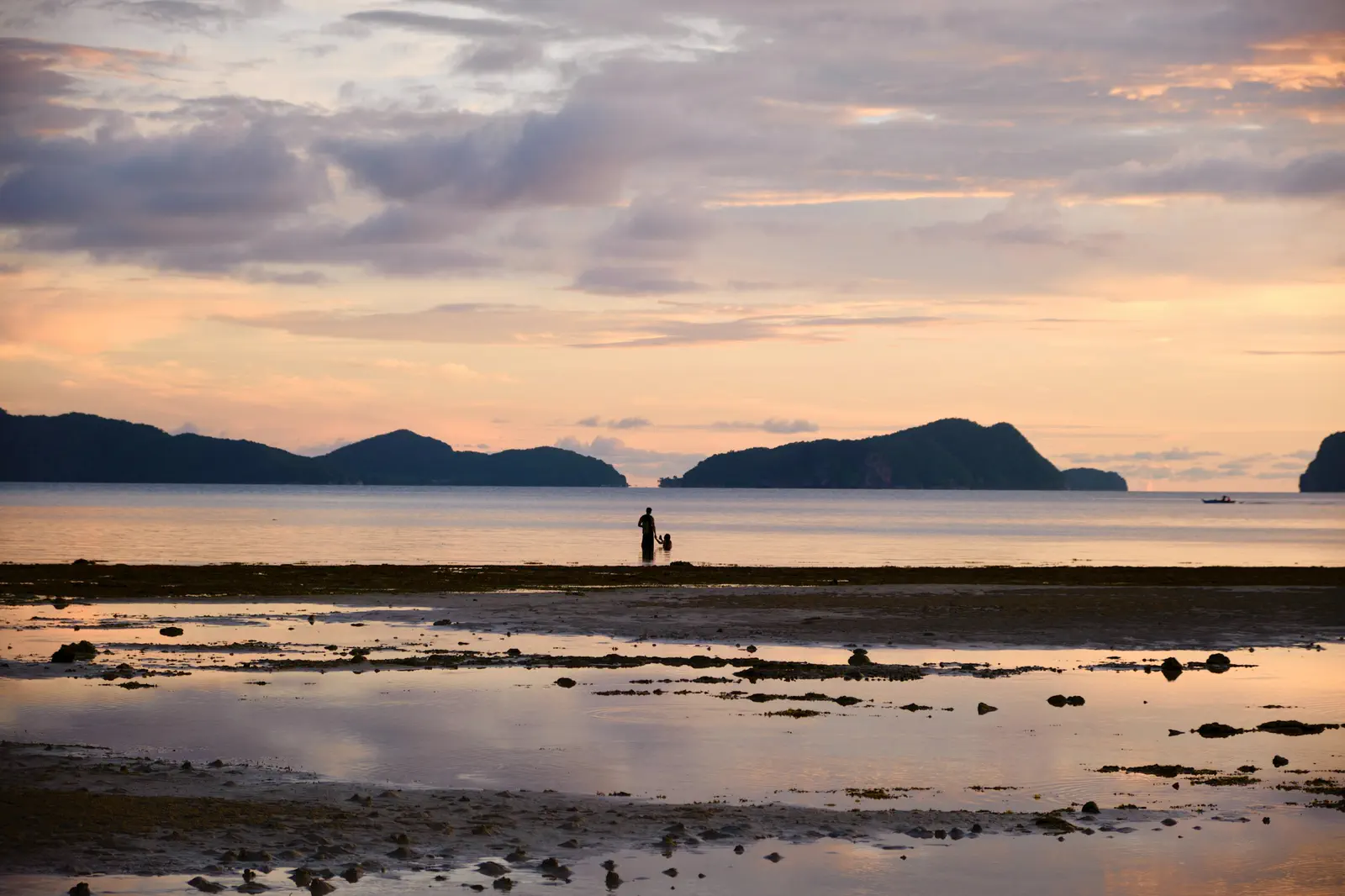 Low tide at dusk in El Nido, Palawan: a parent and child wade far out under a pastel sky between dark island silhouettes