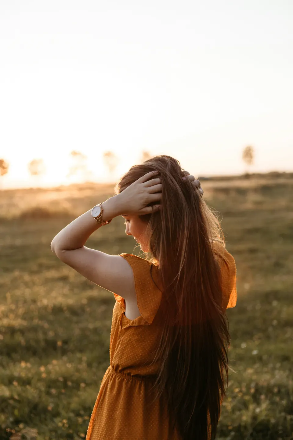A woman in a mustard dress with her hands in her hair, backlit by golden light, face turned away