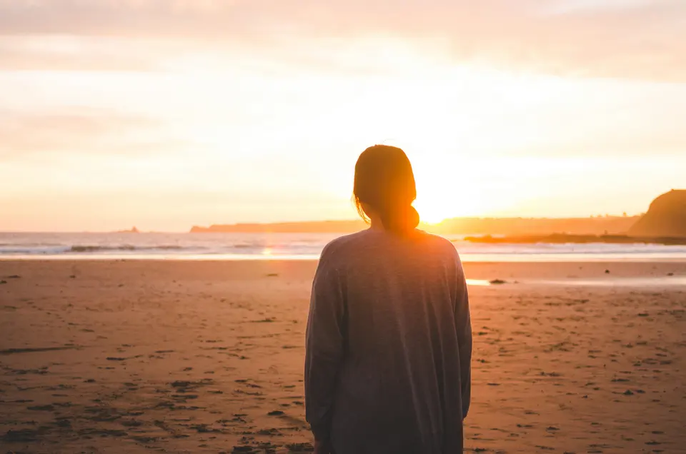 A woman seen from behind on a beach, facing a low setting sun