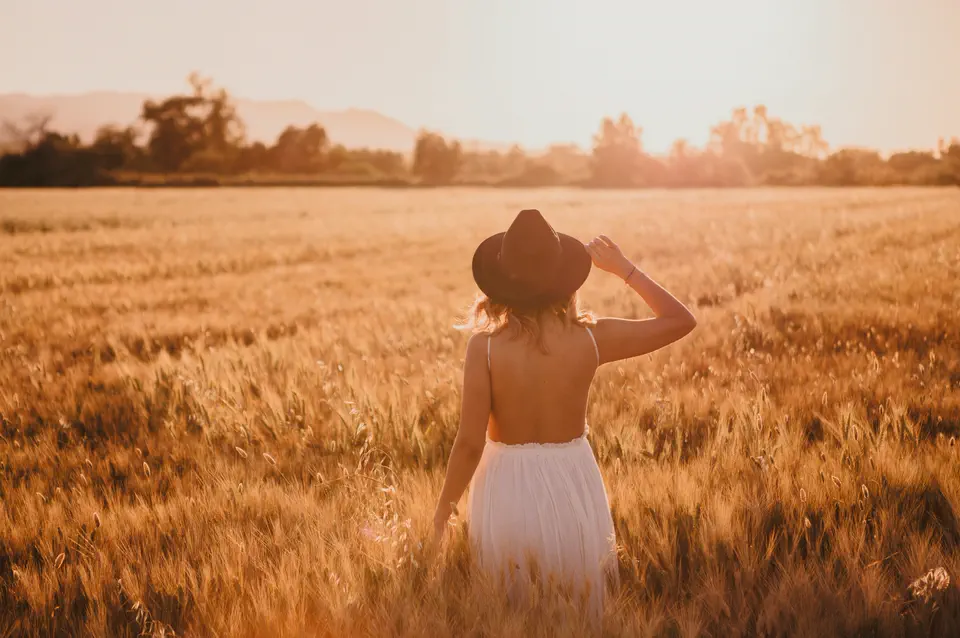 A woman from behind in a golden wheat field, one hand on her hat