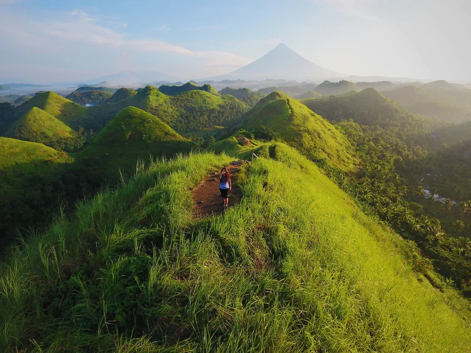 A hiker on a grassy ridge with a cone-shaped volcano on the horizon
