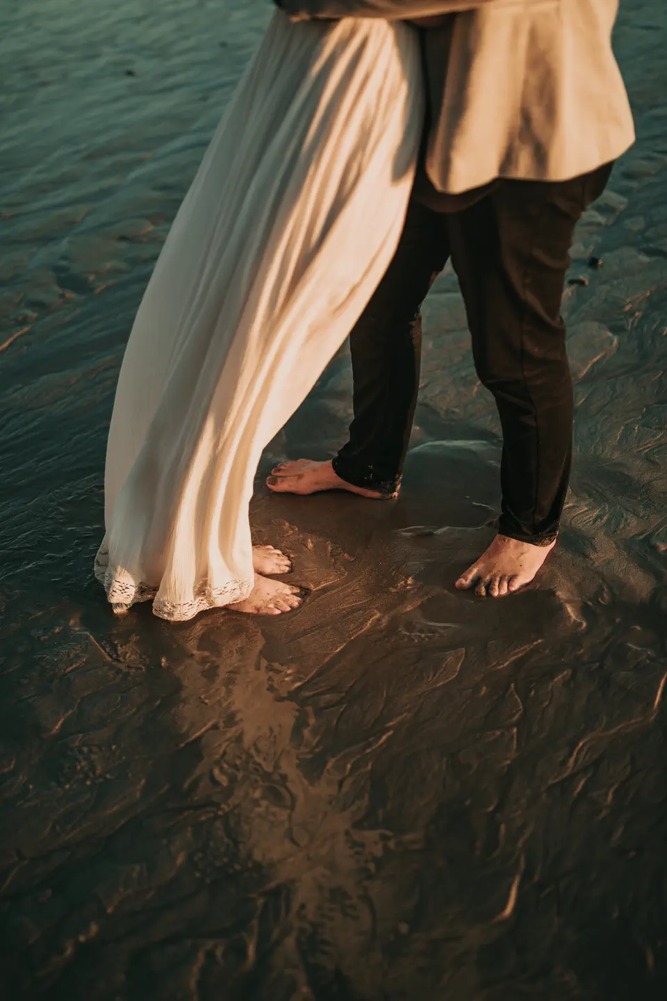 A couple's bare feet in wet sand, the hem of a wedding dress trailing