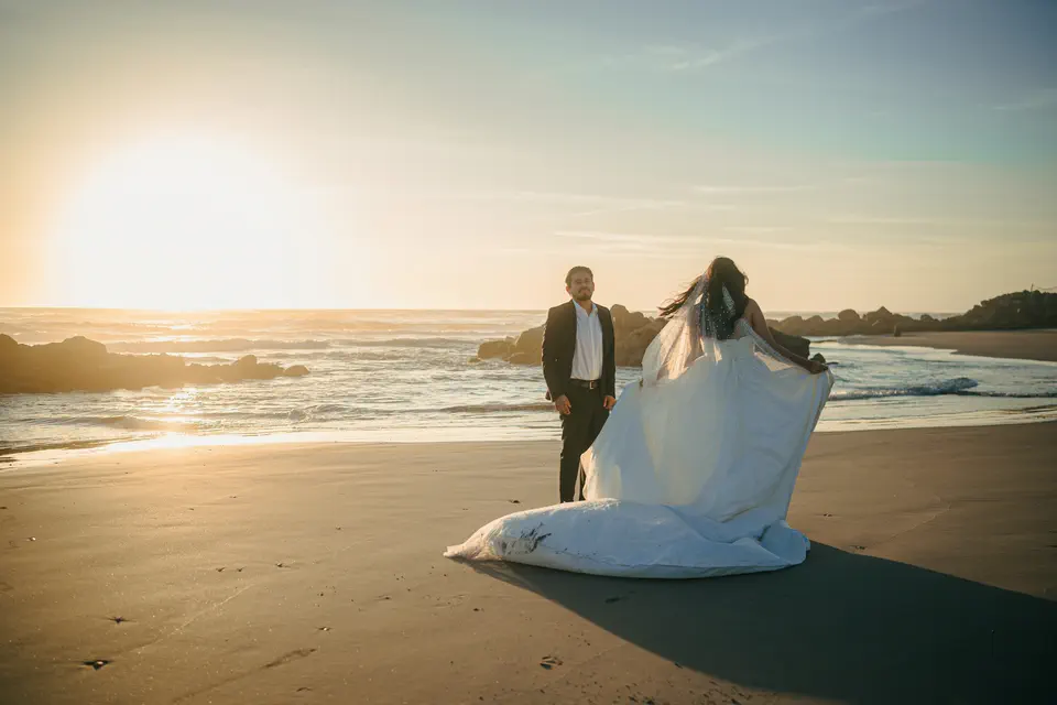 A newly married couple on a wide beach at sunset, her dress lifting in the wind