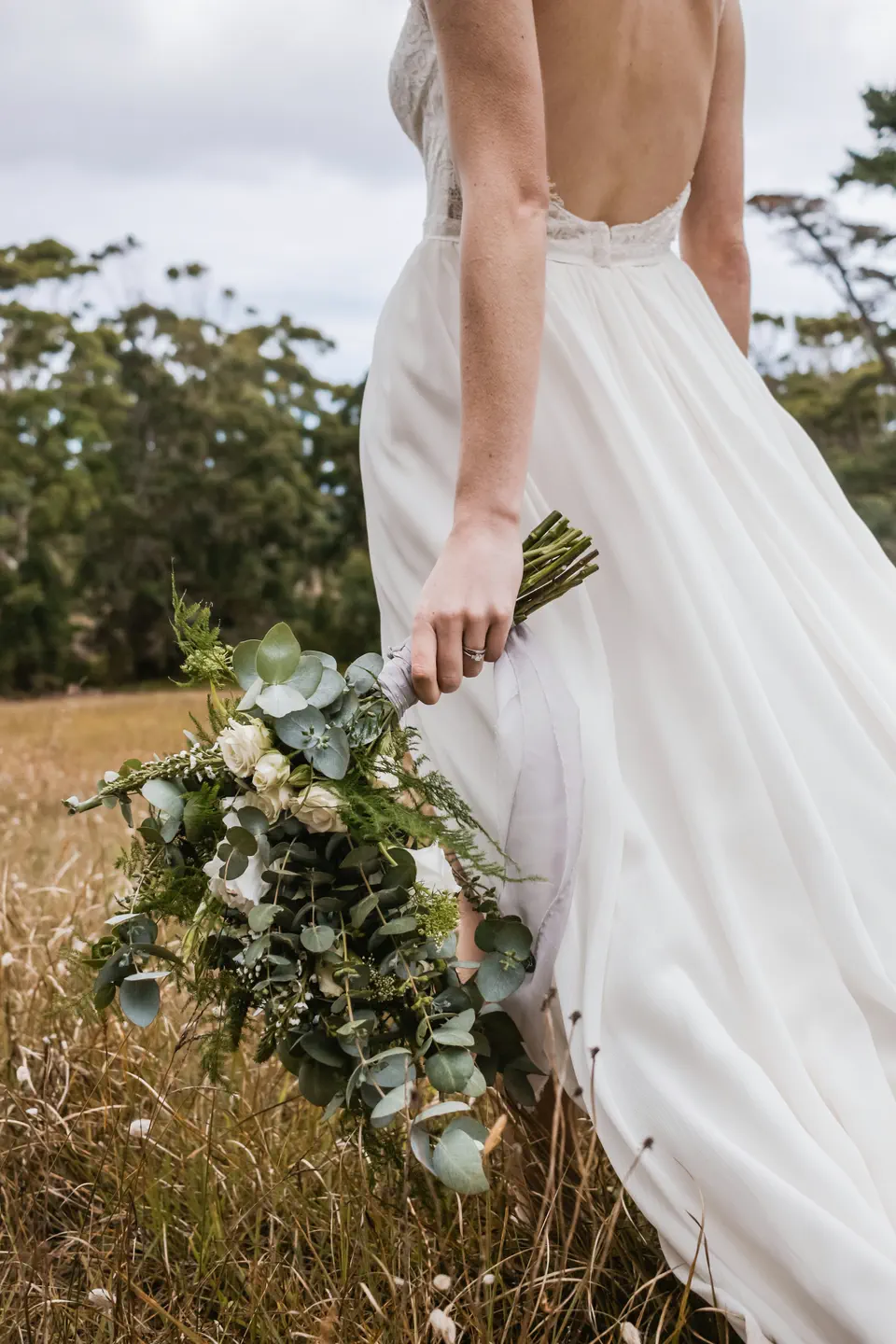 The bride walking through tall grass holding a loose eucalyptus bouquet