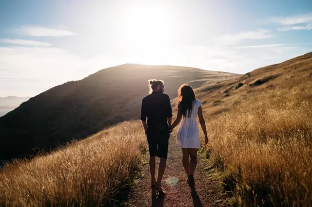 A couple walking hand in hand up a golden, grassy hill at sunrise