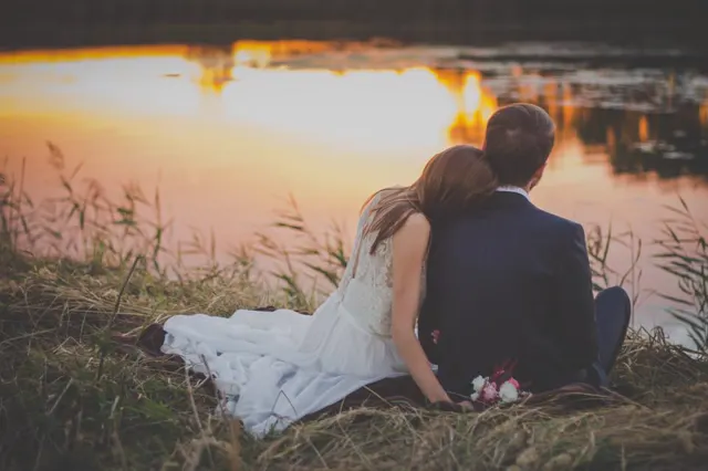 A bride and groom sitting by a lake at sunset, seen from behind