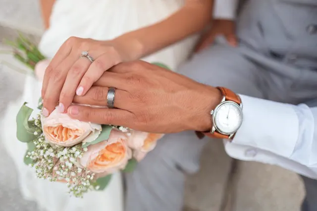 Close-up of two hands with wedding rings resting on a peach rose bouquet