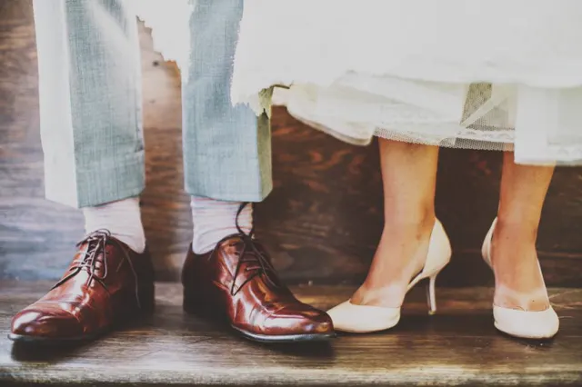The groom's brown shoes and the bride's white heels side by side on a wooden floor